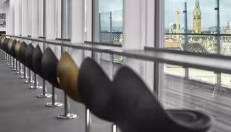 Eine Reihe moderner schwarzer Stühle säumt eine Glaswand in einem stilvollen Interieur. Durch die Fenster ist eine Stadtlandschaft mit historischen Türmen und Gebäuden unter einem teilweise bewölkten Himmel zu sehen.