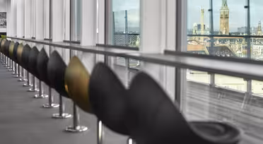 Eine Reihe moderner schwarzer Stühle säumt eine Glaswand in einem stilvollen Interieur. Durch die Fenster ist eine Stadtlandschaft mit historischen Türmen und Gebäuden unter einem teilweise bewölkten Himmel zu sehen.