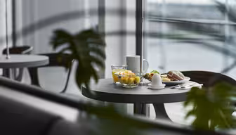 Ein runder Tisch mit Frühstücksgedeck am Fenster: eine Obstschale, ein weichgekochtes Ei, ein Brötchen, ein Glas Wasser und eine Tasse. Im Vordergrund sind unscharfe grüne Blätter zu sehen, und draußen vor dem Fenster sind verschwommene Gebäude zu sehen.