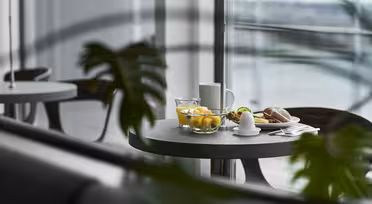 Ein runder Tisch mit Frühstücksgedeck am Fenster: eine Obstschale, ein weichgekochtes Ei, ein Brötchen, ein Glas Wasser und eine Tasse. Im Vordergrund sind unscharfe grüne Blätter zu sehen, und draußen vor dem Fenster sind verschwommene Gebäude zu sehen.