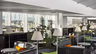 Moderne Lounge mit runden Tischen, grauen Stühlen und grünen und orangefarbenen Kissen. Weiße Lampen auf jedem Tisch, Pflanzen als Dekoration. Große Fenster bieten einen Blick auf die Stadt im Hintergrund. Helle, luftige Atmosphäre.