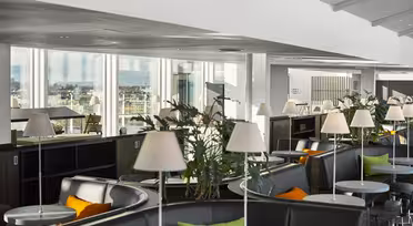 Moderne Lounge mit runden Tischen, grauen Stühlen und grünen und orangefarbenen Kissen. Weiße Lampen auf jedem Tisch, Pflanzen als Dekoration. Große Fenster bieten einen Blick auf die Stadt im Hintergrund. Helle, luftige Atmosphäre.