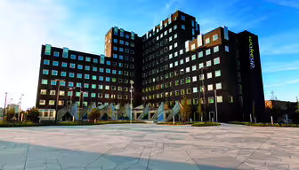 Modernes, mehrstöckiges schwarzes Gebäude mit einzigartigem, kantigem Design und zahlreichen quadratischen Fenstern. Das Gebäude steht vor einem strahlend blauen Himmel und ist von einem großzügigen, geometrisch gemusterten Platz und minimalistischer Landschaftsgestaltung umgeben.