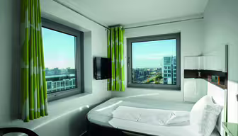Hotelzimmer mit modernem Design, einem ordentlich gemachten Bett, zwei großen Fenstern mit grünen und weißen Vorhängen, einem kleinen an der Wand montierten Fernseher und einem Blick auf eine Stadtlandschaft mit Gebäuden und Straßen in der Ferne.