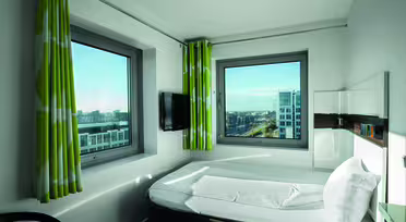 Hotelzimmer mit modernem Design, einem ordentlich gemachten Bett, zwei großen Fenstern mit grünen und weißen Vorhängen, einem kleinen an der Wand montierten Fernseher und einem Blick auf eine Stadtlandschaft mit Gebäuden und Straßen in der Ferne.