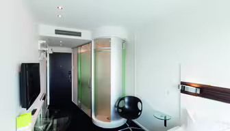 Modernes Hotelzimmer mit runder Glasdusche, schwarzem Stuhl, kleinem Glastisch und Wandfernseher. Das Zimmer ist minimalistisch mit weißen Wänden und schwarzem Boden. Rechts ist ein Bett mit weißer Bettwäsche teilweise sichtbar.