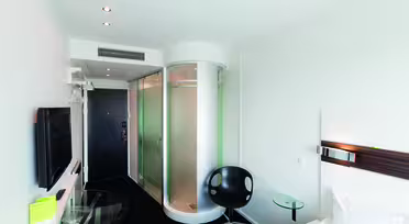 Modernes Hotelzimmer mit runder Glasdusche, schwarzem Stuhl, kleinem Glastisch und Wandfernseher. Das Zimmer ist minimalistisch mit weißen Wänden und schwarzem Boden. Rechts ist ein Bett mit weißer Bettwäsche teilweise sichtbar.