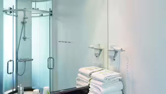 Modernes Badezimmer mit schwarzer Granitplatte und weißem Waschbecken. Handtücher liegen auf der Ablage neben einem wandmontierten Haartrockner. Ein großer Spiegel spiegelt den Duschbereich mit Glastüren wider. Deckeneinbauleuchten erhellen den Raum.