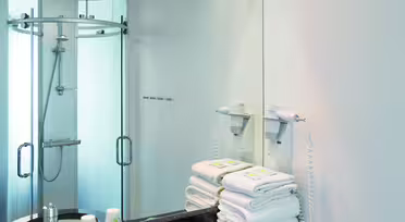 Modernes Badezimmer mit schwarzer Granitplatte und weißem Waschbecken. Handtücher liegen auf der Ablage neben einem wandmontierten Haartrockner. Ein großer Spiegel spiegelt den Duschbereich mit Glastüren wider. Deckeneinbauleuchten erhellen den Raum.
