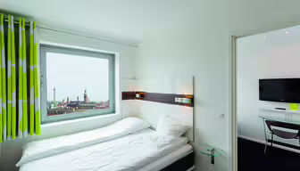 Ein helles Hotelzimmer mit einem ordentlich gemachten Bett und einem Fenster mit Stadtblick. Das Fenster ist mit grün-weiß gestreiften Vorhängen versehen. Im angrenzenden Zimmer hängt ein Flachbildfernseher an der Wand über einem kleinen Tisch.