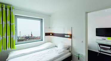 Ein helles Hotelzimmer mit einem ordentlich gemachten Bett und einem Fenster mit Stadtblick. Das Fenster ist mit grün-weiß gestreiften Vorhängen versehen. Im angrenzenden Zimmer hängt ein Flachbildfernseher an der Wand über einem kleinen Tisch.
