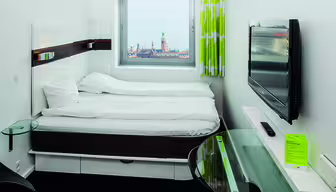 Modernes Hotelzimmer mit Doppelbett und gegenüberliegendem Wandfernseher. Darunter befindet sich ein kleiner Glasschreibtisch. Durch ein Fenster ist eine Stadtlandschaft mit einem Turm in der Ferne zu sehen. Das Zimmer hat weiße Wände und einen hellgrünen Vorhang.