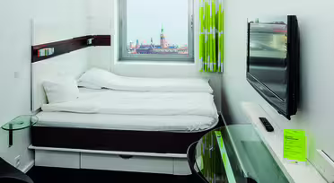 Modernes Hotelzimmer mit Doppelbett und gegenüberliegendem Wandfernseher. Darunter befindet sich ein kleiner Glasschreibtisch. Durch ein Fenster ist eine Stadtlandschaft mit einem Turm in der Ferne zu sehen. Das Zimmer hat weiße Wände und einen hellgrünen Vorhang.