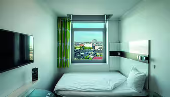 Ein kleines, modernes Hotelzimmer mit einem Einzelbett am Fenster. Das Fenster bietet einen Blick auf die Stadt. Das Zimmer verfügt über einen wandmontierten Flachbildfernseher, einen eleganten Schreibtisch und grün gemusterte Vorhänge. Die Einrichtung ist minimalistisch und überwiegend in Weiß gehalten.