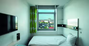 Ein kleines, modernes Hotelzimmer mit einem Einzelbett am Fenster. Das Fenster bietet einen Blick auf die Stadt. Das Zimmer verfügt über einen wandmontierten Flachbildfernseher, einen eleganten Schreibtisch und grün gemusterte Vorhänge. Die Einrichtung ist minimalistisch und überwiegend in Weiß gehalten.