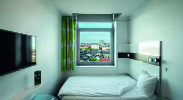 Ein kleines, modernes Hotelzimmer mit einem Einzelbett am Fenster. Das Fenster bietet einen Blick auf die Stadt. Das Zimmer verfügt über einen wandmontierten Flachbildfernseher, einen eleganten Schreibtisch und grün gemusterte Vorhänge. Die Einrichtung ist minimalistisch und überwiegend in Weiß gehalten.