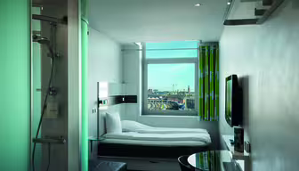 Ein modernes, kompaktes Hotelzimmer mit Einzelbett, Wandfernseher und rundem Tisch. Eine verglaste Dusche befindet sich in der Nähe des Eingangs. Das Fenster bietet einen Blick auf die Stadt mit Gebäuden und Kränen in der Ferne.