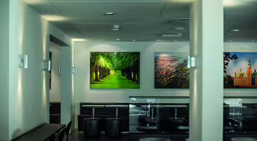 Ein modernes Café mit schwarzen Stühlen und Tischen und großen gerahmten Landschaftsfotos an den Wänden. Das zentrale Foto zeigt einen sonnenbeschienenen, von Bäumen gesäumten Weg. Sanftes Licht und minimalistisches Dekor schaffen eine ruhige Atmosphäre.