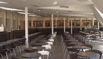 Moderne Cafeteria mit Reihen runder Tische und Holzstühlen. Große Fenster lassen Tageslicht in den großzügigen Raum mit Theke und Deckenleuchten. Die minimalistische Einrichtung schafft eine offene und einladende Atmosphäre.