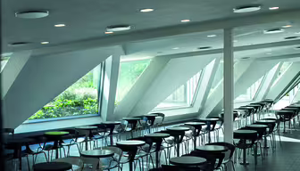 Geräumige, moderne Cafeteria mit Reihen schwarzer Stühle und runden Tischen. Große, schräge Fenster an der Seite lassen viel Tageslicht herein und erhellen das elegante Innendesign.
