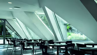 Ein modernes, luftiges Café mit eleganten schwarzen Tischen und Stühlen, das durch große, schräge Fenster mit natürlichem Licht erhellt wird. Die geometrische Architektur schafft eine großzügige und helle Atmosphäre, von der aus man das Grün der Umgebung bewundern kann.