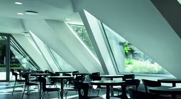 Ein modernes, luftiges Café mit eleganten schwarzen Tischen und Stühlen, das durch große, schräge Fenster mit natürlichem Licht erhellt wird. Die geometrische Architektur schafft eine großzügige und helle Atmosphäre, von der aus man das Grün der Umgebung bewundern kann.