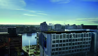 Eine Stadtansicht mit modernen Gebäuden an einem Gewässer unter klarem blauen Himmel. Im Vordergrund ist ein weißes, mehrstöckiges Gebäude zu sehen, im Hintergrund ein Mix verschiedener Architekturstile mit einem fernen Horizont.