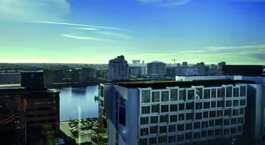 Eine Stadtansicht mit modernen Gebäuden an einem Gewässer unter klarem blauen Himmel. Im Vordergrund ist ein weißes, mehrstöckiges Gebäude zu sehen, im Hintergrund ein Mix verschiedener Architekturstile mit einem fernen Horizont.