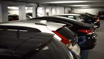 Eine Reihe parkender Autos in einem schwach beleuchteten Parkhaus. Die Fahrzeuge stehen dicht beieinander, die meisten sind dunkel lackiert. Das Parkhaus verfügt über Deckenbeleuchtung und Betonpfeiler.