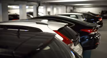 Eine Reihe parkender Autos in einem schwach beleuchteten Parkhaus. Die Fahrzeuge stehen dicht beieinander, die meisten sind dunkel lackiert. Das Parkhaus verfügt über Deckenbeleuchtung und Betonpfeiler.