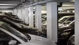 Ein schwach beleuchtetes Parkhaus voller geparkter Autos. Die Fahrzeuge sind überwiegend dunkel lackiert, und das Parkhaus wird von weißen Säulen getragen. Die Szene wirkt überladen und vermittelt den Eindruck eines überfüllten Parkplatzes.