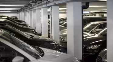 Ein schwach beleuchtetes Parkhaus voller geparkter Autos. Die Fahrzeuge sind überwiegend dunkel lackiert, und das Parkhaus wird von weißen Säulen getragen. Die Szene wirkt überladen und vermittelt den Eindruck eines überfüllten Parkplatzes.