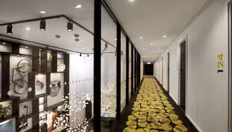 Ein moderner Hotelflur mit eleganten Designelementen. Die rechte Seite ist mit weißen Türen, darunter 124, versehen, während die linke Seite eine Glaswand mit dekorativen Regalen und Kunstwerken beherbergt. Der Teppich ist mit kreisförmigen gelben Mustern verziert.