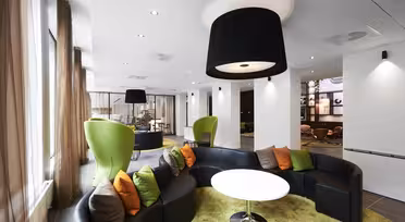 Moderner Loungebereich mit geschwungenen schwarzen Sofas und grünen Sesseln um runde weiße Tische. Der Boden ist mit grünem Teppich ausgelegt, und darüber hängen übergroße schwarze Deckenleuchten. Große Fenster mit transparenten Vorhängen säumen eine Seite und sorgen für natürliches Licht.