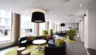 Moderne Lounge mit schwarzen Rundsofas, grünen Sesseln und runden Tischen auf limettengrünen Teppichen. Große Fenster und Deckenleuchten erhellen den Raum. Draußen ist durch transparente Vorhänge eine Stadtstraße sichtbar.
