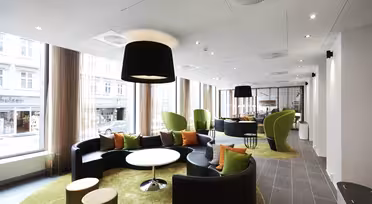 Moderne Lounge mit schwarzen Rundsofas, grünen Sesseln und runden Tischen auf limettengrünen Teppichen. Große Fenster und Deckenleuchten erhellen den Raum. Draußen ist durch transparente Vorhänge eine Stadtstraße sichtbar.