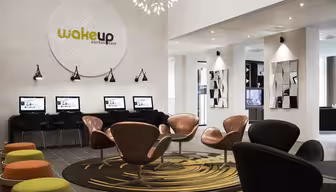 Moderne Hotellobby mit elegantem Design. Mehrere Computerstationen befinden sich an der Wand unter einem großen runden Logo mit der Aufschrift „Wakeup“. Plüschdrehstühle und bunte Hocker stehen auf gemusterten Teppichen. Von der Decke hängt eine geometrische Leuchte.