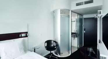 Modernes Hotelzimmer mit minimalistischem Design. Es verfügt über ein Bett mit weißer Bettwäsche, einen schwarzen Stuhl, einen runden Glastisch und eine einzigartige zylindrische Glasdusche in der Nähe des Eingangs. An der Wand gegenüber dem Bett ist ein Flachbildfernseher angebracht.