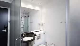 Modernes Hotelbadezimmer mit Glastür. Über einer schwarzen Waschtischplatte befindet sich ein großer Spiegel mit Waschbecken, Haartrockner und gefalteten Handtüchern. Neben der Waschtischplatte befindet sich eine Toilette, in der Ecke eine verglaste Dusche. Schlichtes und elegantes Design.