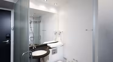 Modernes Hotelbadezimmer mit Glastür. Über einer schwarzen Waschtischplatte befindet sich ein großer Spiegel mit Waschbecken, Haartrockner und gefalteten Handtüchern. Neben der Waschtischplatte befindet sich eine Toilette, in der Ecke eine verglaste Dusche. Schlichtes und elegantes Design.