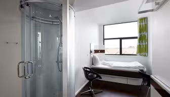 Kompaktes, modernes Hotelzimmer mit einem Einzelbett an einem großen Fenster, einem kleinen Schreibtisch mit schwarzem Stuhl und einer runden Glasduschkabine in einer Ecke. Das Zimmer verfügt über dunklen Holzboden und grün gemusterte Vorhänge.