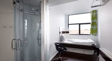 Kompaktes, modernes Hotelzimmer mit einem Einzelbett an einem großen Fenster, einem kleinen Schreibtisch mit schwarzem Stuhl und einer runden Glasduschkabine in einer Ecke. Das Zimmer verfügt über dunklen Holzboden und grün gemusterte Vorhänge.