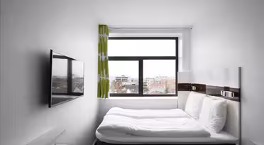 Ein kompaktes Hotelzimmer mit weißen Wänden und einem Doppelbett mit weißer Bettwäsche. An der linken Wand hängt ein Flachbildfernseher. Ein großes Fenster mit grüngestreiftem Vorhang bietet einen Blick auf die Stadt. Minimalistisches Dekor wertet den Raum auf.