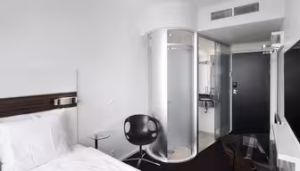 Modernes Hotelzimmer mit minimalistischem Design, ausgestattet mit weißem Bett, schwarzem Stuhl, Glasschreibtisch und zylindrischer Badezimmerkabine aus Milchglas. Wände und Bettwäsche sind weiß und bilden einen Kontrast zum dunklen Boden und den Möbeln.