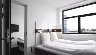 Ein modernes, minimalistisches Hotelzimmer verfügt über ein ordentlich gemachtes Bett mit weißer Bettwäsche, einen Nachttisch aus Glas und ein Fenster mit Stadtblick. Eine Tür führt zu einem angrenzenden Zimmer mit einem weiteren Bett und einem an der Wand montierten Fernseher.