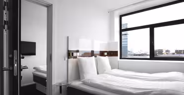 Ein modernes, minimalistisches Hotelzimmer verfügt über ein ordentlich gemachtes Bett mit weißer Bettwäsche, einen Nachttisch aus Glas und ein Fenster mit Stadtblick. Eine Tür führt zu einem angrenzenden Zimmer mit einem weiteren Bett und einem an der Wand montierten Fernseher.