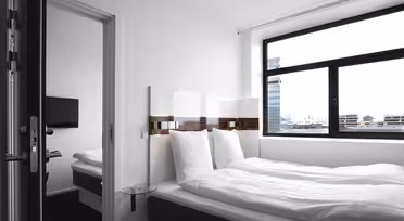 Ein modernes, minimalistisches Hotelzimmer verfügt über ein ordentlich gemachtes Bett mit weißer Bettwäsche, einen Nachttisch aus Glas und ein Fenster mit Stadtblick. Eine Tür führt zu einem angrenzenden Zimmer mit einem weiteren Bett und einem an der Wand montierten Fernseher.
