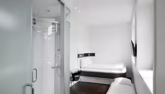 Minimalistisches Hotelzimmer mit kleiner, geschlossener Dusche mit Milchglastüren. Es gibt ein Bett, einen an der Wand montierten Fernseher, einen Schreibtisch mit Stuhl und ein schmales Fenster auf der rechten Seite. Der Raum ist modern und in neutralen Weiß- und Holztönen gehalten.