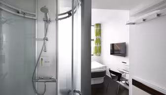 Ein kompaktes Hotelzimmer verfügt über eine moderne Glasdusche, ein Bett, einen wandmontierten Fernseher, einen Schreibtisch und einen Stuhl. Die Einrichtung ist schlicht und elegant, mit einem grün gemusterten Vorhang und weißen Wänden, die das helle, minimalistische Design unterstreichen.