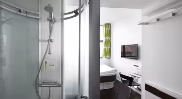 Ein kompaktes Hotelzimmer verfügt über eine moderne Glasdusche, ein Bett, einen wandmontierten Fernseher, einen Schreibtisch und einen Stuhl. Die Einrichtung ist schlicht und elegant, mit einem grün gemusterten Vorhang und weißen Wänden, die das helle, minimalistische Design unterstreichen.